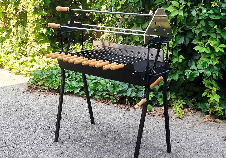 A black metal outdoor barbecue grill with wooden handles and a stainless steel rotisserie rod, set on a paved surface with green foliage in the background.